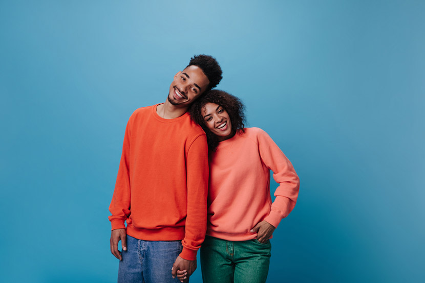 Couple in love looking into camera on blue background. Portrait of happy curly woman in orange sweater and dark-skinned man in jeans smiling on isolated