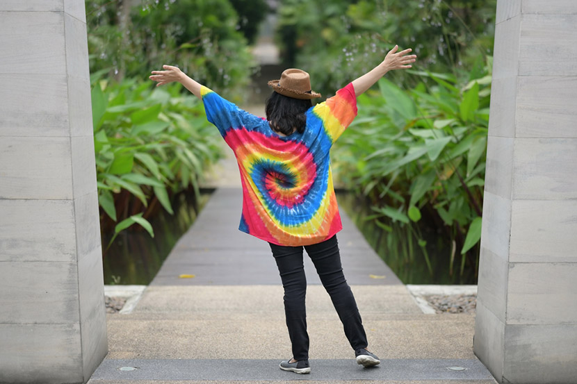 A woman Wear a bright tie-dye shirt Wearing a brown straw hat and black jeans stand with your back raises both her hands above her head while walking on  wooden bridge to admire beauty of green garden